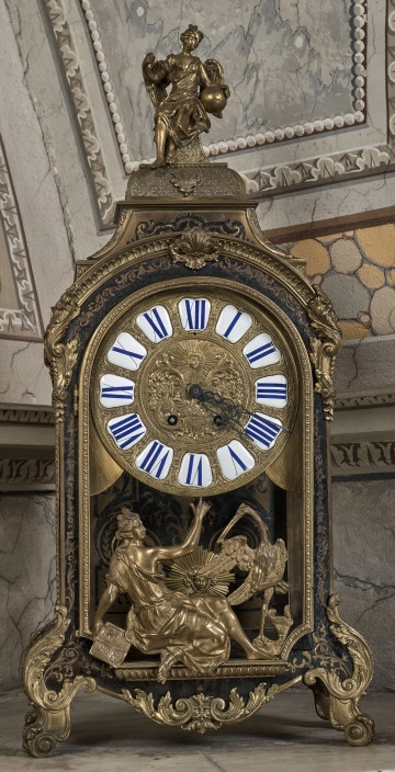 Przód stojącego zegara z mosiężnymi inkrustacjami i złoceniami. W części centralnej złota tarcza z cyframi rzymskimi. W części dolnej i w szczycie przedstawienia figuralne.