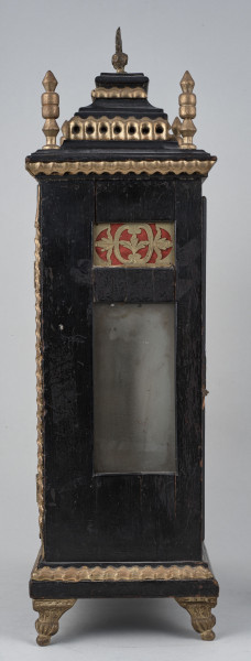 Zegar szafkowy z uchwytem górnym do przenoszenia. Prostopadłościenna obudowa drewniana, czerniona, złocona, posiada aplikacje mosiężne. Ścianka boczna z otworem, przez który widać mechanizm.