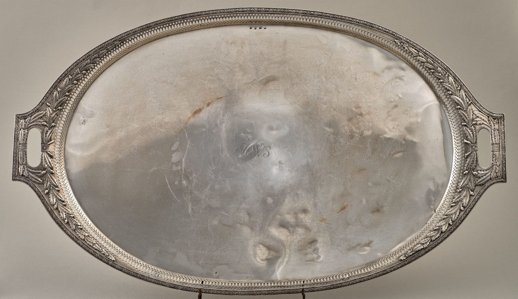 Taca srebrna, owalna z dekoracyjnym brzegiem z dwoma uchwytami ustawiona na drucianej podstawce.