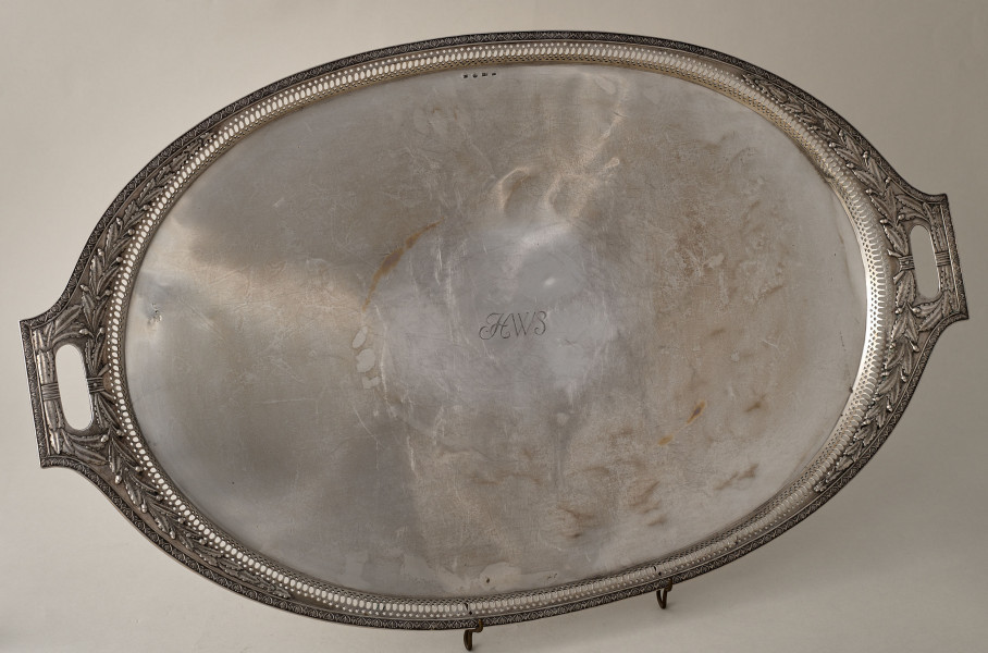 Taca srebrna, owalna z dekoracyjnym brzegiem z dwoma uchwytami ustawiona na drucianej podstawce.
