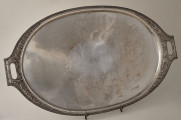 Taca srebrna, owalna z dekoracyjnym brzegiem z dwoma uchwytami ustawiona na drucianej podstawce.