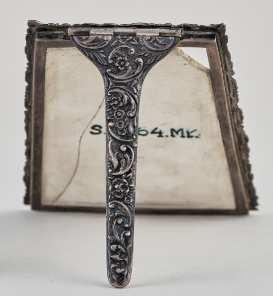 Ramka na menu. Ramka srebrna dekorowana ornamentem roślinnym.Wewnątrz płytka ceramiczna, pęknięta pionowo. Widok od tyłu z widoczną podpórką.