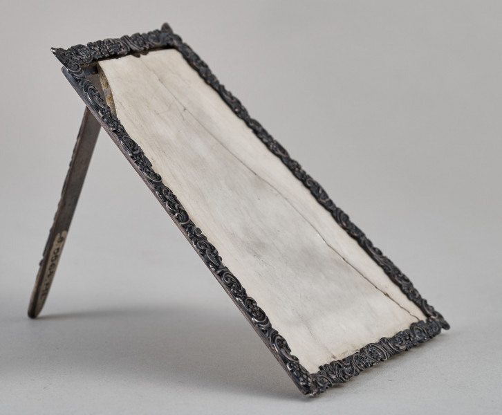 Ramka na menu. Ramka srebrna dekorowana ornamentem roślinnym.Wewnątrz płytka ceramiczna, pęknięta pionowo. Ujęcie boczne z widoczną podpórką.