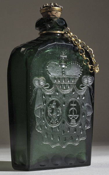 Flasza z zielonego szkła z herbem Trąby i Waga. Widoczny złoty łańcuszek z korkiem.