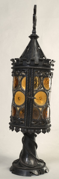 Latarnia w kształcie cylindra zwieńczonego stożkowym metalowym nakryciem, ażurowo wycinanym, zakończonym sterczyną w kształcie zbliżonym do lilii andegaweńskiej. Stopa latarni okrągła o fantazyjnie wycinanym brzegu, ozdobiona jest ornamentem perełkowym w kształcie oplatającego ją sznura. Całość obudowy (cylindra) latarni zdobi witraż (szkło w odcieniach żółci), ujęty w metalowe obramienia zdobione ornamentem perełkowym.