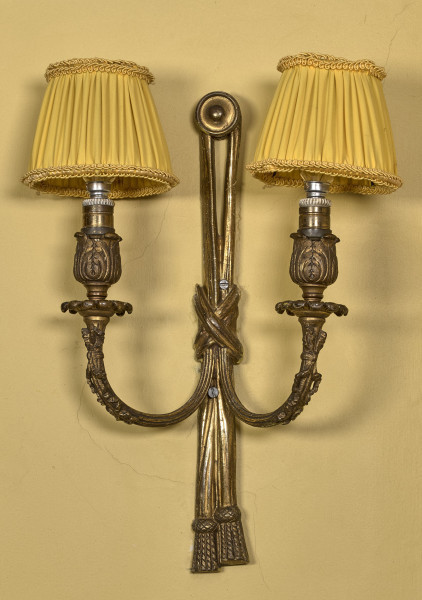 Świecznik przyścienny - aplika. Trzon w formie wąskiego kawałka tkaniny przełożonej przez ozdobny kołek – rozetę przewiązany pośrodku skrzyżowaną wstęgą. Dołem oba kawałki tkaniny zakończone są chwostem z frędzlami. Żarówki osłonięte żółtymi abażurkami. Aplika na tle żółtej ściany.