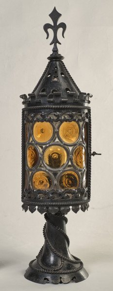 Latarnia w kształcie cylindra zwieńczonego stożkowym metalowym nakryciem, ażurowo wycinanym, zakończonym sterczyną w kształcie zbliżonym do lilii andegaweńskiej. Stopa latarni okrągła o fantazyjnie wycinanym brzegu, ozdobiona jest ornamentem perełkowym w kształcie oplatającego ją sznura. Całość obudowy (cylindra) latarni zdobi witraż (szkło w odcieniach żółci), ujęty w metalowe obramienia zdobione ornamentem perełkowym.