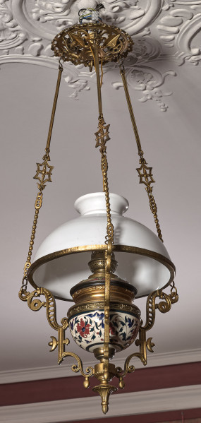 Lampa wisząca na tle sufitu i ściany. Lampa na długich łańcuszkach z białym kloszem z mosiężnymi okuciami.