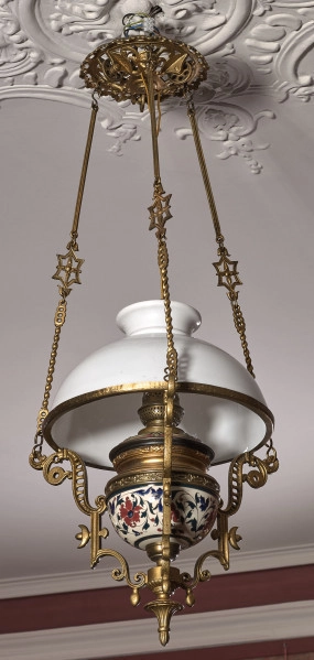 Lampa wisząca na tle sufitu i ściany. Lampa na długich łańcuszkach z białym kloszem z mosiężnymi okuciami.