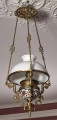 Lampa wisząca na tle sufitu i ściany. Lampa na długich łańcuszkach z białym kloszem z mosiężnymi okuciami.