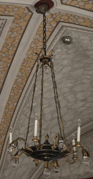 Lampa wisząca na łańcuchach.  Korpus, malowany na czarno, w kształcie stożkowatego dysku, do którego przymocowano dziesięć punktów świetlnych czyli pięć ramion wygiętych esowato ku górze, zakończonych tulejkami drewnianymi malowanymi na biało zakończonych żarówkami świecowymi,  a pięć innych wygiętych ku dołowi w kształcie gałązki zakończonej kwiatonem z umieszczonymi w środku żarówkami. W tle widaćsufit dekorowany malarską imitacją ściany kamiennej.