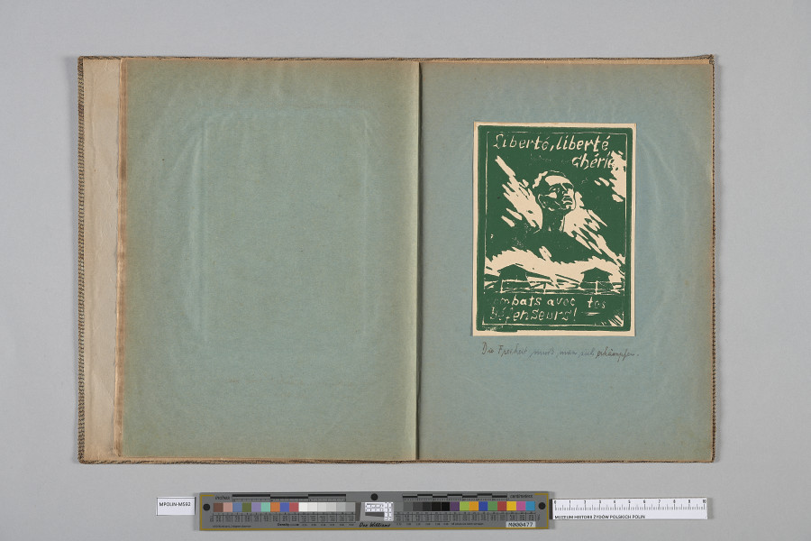 Ujęcie od góry. Rozłożony zeszyt z grafiką naklejoną na prawą kartę. Grafika przedstawia  głowe mężczyzny nad dwoma, mniejszymi, barakami i płotem.