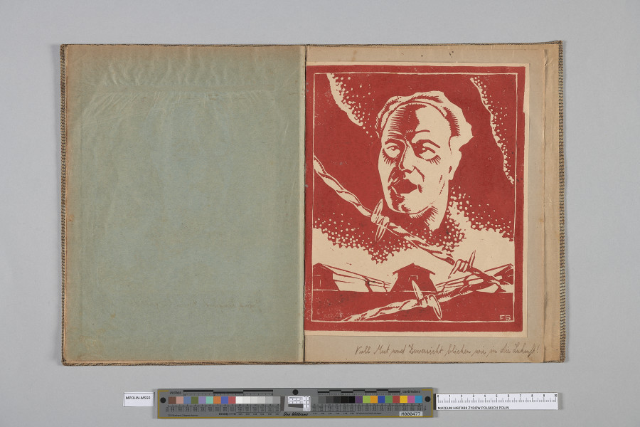 Ujęcie od góry. Rozłożony zeszyt z grafiką naklejoną na prawą kartę. Grafika przedstawia  głowę mężczyzny ujętą w trzech czwartych na tle dekoracyjnych chmur; niżej przecinające się na pierwszym planie fragmenty drutu kolczastego.