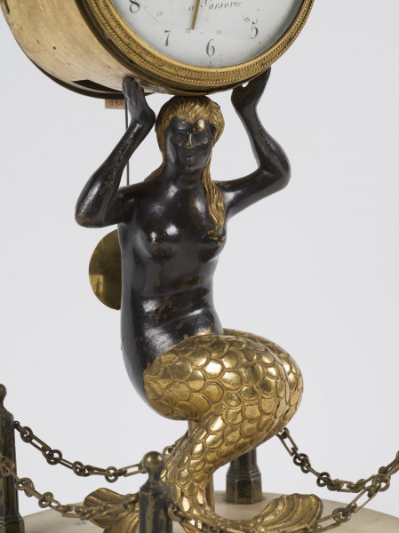 zbliżenie na postać Syreny podtrzymującą mechanizm zegara z okrągłą tarczą - włosy oraz ogon Syreny złocone, pozostała część lakierowana na czarno