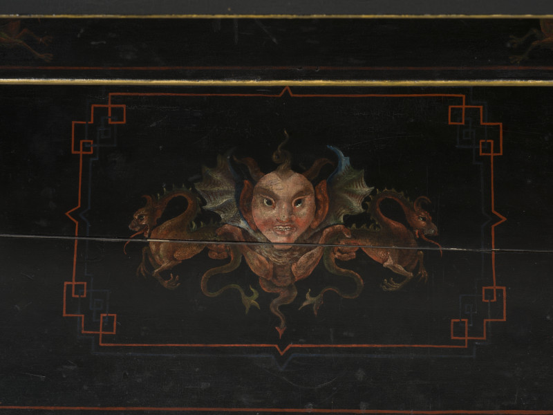 zbliżenie na blat biurka zdobiony wielobarwną dekoracją malarską, przedstawiającą dużą uskrzydloną maskę ujętą w dwa smoki ustawione od siebie