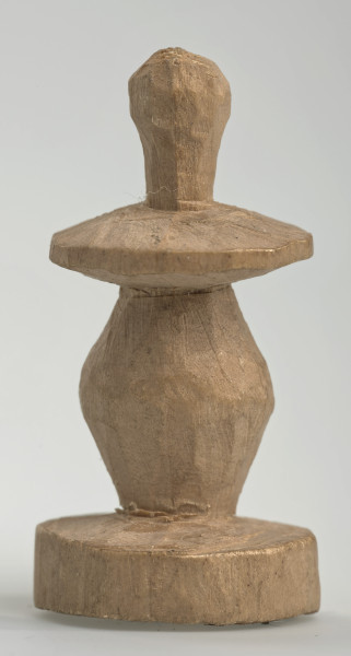 Ujęcie z boku [3]. Ze względu na swój okrągły kształt kolejne ujęcia z boku nie różnią się między sobą znacząco. Pionek został wykonany ręcznie z jednego kawałka jasnego drewna. Na całej powierzchni widoczne są ślady rzeźbienia przy pomocy noża. Obiekt w kolorze naturalnego drewna (nie został pomalowany, ani polakierowany). Od spodu widoczne oznaczenie składające się z dwóch przecinających się ze sobą linii („X”), które umożliwia podzielenie bierek pomiędzy dwóch graczy - wszystkie bierki są jednakowego koloru. Krawędź podstawy została dodatkowo obrysowana ołówkiem. Pionek składa się ze spłaszczonej podstawy w formie krążka, części środkowej w kształcie gruszki, spłaszczonego ronda, które zwęża się stopniowo ku górze oraz ku dołowi (przypominając dwa połączone ze sobą talerze, z których każdy skierowany jest dnem do zewnątrz) oraz zwieńczenia o prostej, rozszerzającej się ku górze formie (przypominającej pałkę).