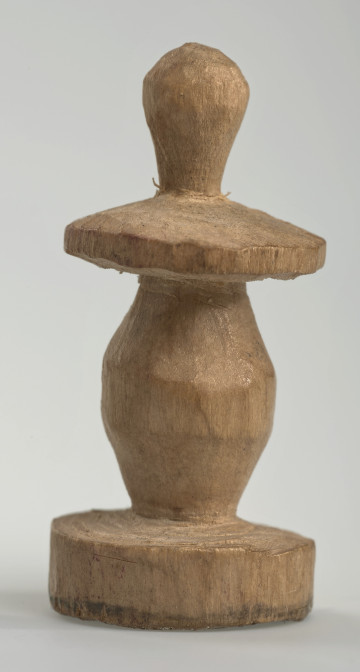 Ujęcie z boku [1]. Ze względu na swój okrągły kształt kolejne ujęcia z boku nie różnią się między sobą znacząco. Pionek został wykonany ręcznie z jednego kawałka jasnego drewna. Na całej powierzchni widoczne są ślady rzeźbienia przy pomocy noża. Obiekt w kolorze naturalnego drewna (nie został pomalowany, ani polakierowany). Krawędź podstawy została dodatkowo obrysowana ołówkiem. Pionek składa się ze spłaszczonej podstawy w formie krążka, części środkowej w kształcie gruszki, spłaszczonego ronda, które zwęża się stopniowo ku górze oraz ku dołowi (przypominając dwa połączone ze sobą talerze, z których każdy skierowany jest dnem do zewnątrz) oraz zwieńczenia o prostej, rozszerzającej się ku górze formie (przypominającej pałkę).