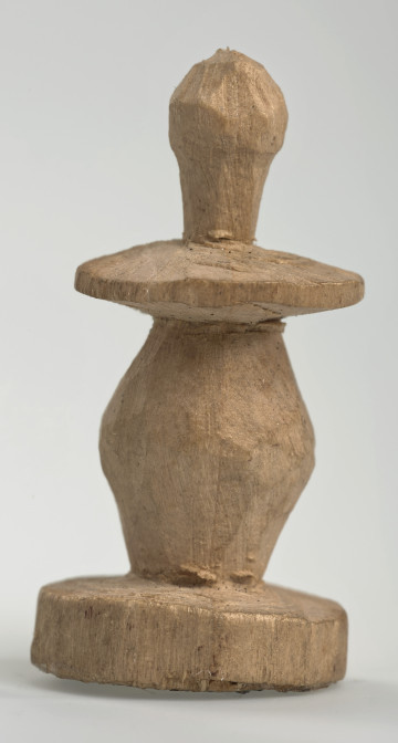 Ujęcie z boku [1]. Ze względu na swój okrągły kształt kolejne ujęcia z boku nie różnią się między sobą znacząco. Pionek został wykonany ręcznie z jednego kawałka jasnego drewna. Na całej powierzchni widoczne są ślady rzeźbienia przy pomocy noża. Obiekt w kolorze naturalnego drewna (nie został pomalowany, ani polakierowany). Od spodu widoczne oznaczenie składające się z dwóch przecinających się ze sobą linii („X”), które umożliwia podzielenie bierek pomiędzy dwóch graczy - wszystkie bierki są jednakowego koloru. Krawędź podstawy została dodatkowo obrysowana ołówkiem. Pionek składa się ze spłaszczonej podstawy w formie krążka, części środkowej w kształcie gruszki, spłaszczonego ronda, które zwęża się stopniowo ku górze oraz ku dołowi (przypominając dwa połączone ze sobą talerze, z których każdy skierowany jest dnem do zewnątrz) oraz zwieńczenia o prostej, rozszerzającej się ku górze formie (przypominającej pałkę).