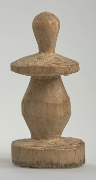 Ujęcie z boku [2]. Ze względu na swój okrągły kształt kolejne ujęcia z boku nie różnią się między sobą znacząco. Pionek został wykonany ręcznie z jednego kawałka jasnego drewna. Na całej powierzchni widoczne są ślady rzeźbienia przy pomocy noża. Obiekt w kolorze naturalnego drewna (nie został pomalowany, ani polakierowany). Krawędź podstawy została dodatkowo obrysowana ołówkiem. Pionek składa się ze spłaszczonej podstawy w formie krążka, części środkowej w kształcie gruszki, spłaszczonego ronda, które zwęża się stopniowo ku górze oraz ku dołowi (przypominając dwa połączone ze sobą talerze, z których każdy skierowany jest dnem do zewnątrz) oraz zwieńczenia o prostej, rozszerzającej się ku górze formie (przypominającej pałkę).