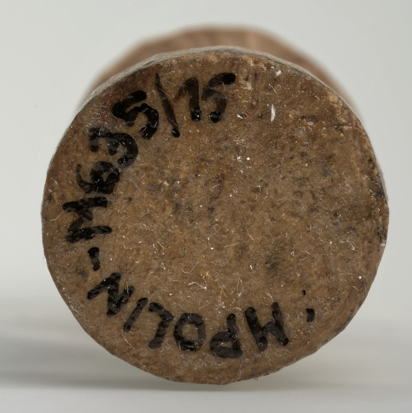 Ujęcie na spód obiektu. Okrągła podstawa pionka szachowego. Ponieważ był on rzeźbiony ręcznie krawędź nie jest idealnie równa. Wzdłuż krawędzi widać czarny numer inwentarzowy MPOLIN-M635/15.