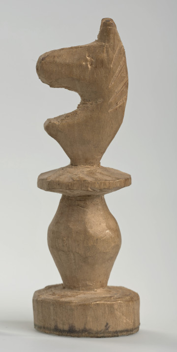 Ujęcie lewego boku. Skoczek został wykonany ręcznie z jednego kawałka jasnego drewna. Na całej powierzchni widoczne są ślady rzeźbienia przy pomocy noża. Obiekt w kolorze naturalnego drewna (nie został pomalowany, ani polakierowany). Od spodu widoczne oznaczenie składające się z dwóch przecinających się ze sobą linii („X”), które umożliwia podzielenie bierek pomiędzy dwóch graczy - wszystkie bierki są jednakowe. Krawędź podstawy została dodatkowo obrysowana ołówkiem. Skoczek składa się ze spłaszczonej podstawy w formie krążka, części środkowej w formie walca rozszerzającego się po środku wysokości, spłaszczonego cylindrycznego elementu, który zwęża się stopniowo ku górze oraz ku dołowi (przypominając dwa połączone ze sobą talerze, z których każdy skierowany jest dnem do zewnątrz) oraz zwieńczenia w formie nawiązującej do głowy i szyi konia. Głowa oraz szyja są lekko spłaszczone. Widoczna wyżłobiona grzywa i oko.