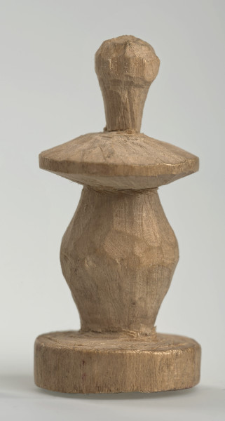 Ujęcie z boku [1]. Ze względu na swój okrągły kształt kolejne ujęcia z boku nie różnią się między sobą znacząco. Pionek został wykonany ręcznie z jednego kawałka jasnego drewna. Na całej powierzchni widoczne są ślady rzeźbienia przy pomocy noża. Obiekt w kolorze naturalnego drewna (nie został pomalowany, ani polakierowany). Krawędź podstawy została dodatkowo obrysowana ołówkiem. Pionek składa się ze spłaszczonej podstawy w formie krążka, części środkowej w kształcie gruszki, spłaszczonego ronda, które zwęża się stopniowo ku górze oraz ku dołowi (przypominając dwa połączone ze sobą talerze, z których każdy skierowany jest dnem do zewnątrz) oraz zwieńczenia o prostej, rozszerzającej się ku górze formie (przypominającej pałkę).