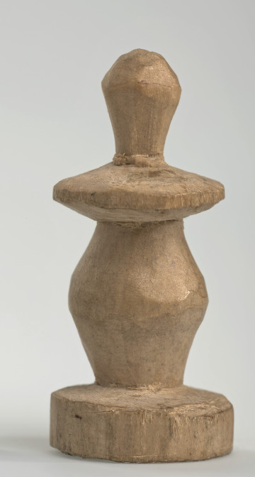 Ujęcie z boku [1]. Ze względu na swój okrągły kształt kolejne ujęcia z boku nie różnią się między sobą znacząco. Pionek został wykonany ręcznie z jednego kawałka jasnego drewna. Na całej powierzchni widoczne są ślady rzeźbienia przy pomocy noża. Obiekt w kolorze naturalnego drewna (nie został pomalowany, ani polakierowany). Od spodu widoczne oznaczenie składające się z dwóch przecinających się ze sobą linii („X”), które umożliwia podzielenie bierek pomiędzy dwóch graczy - wszystkie bierki są jednakowego koloru. Krawędź podstawy została dodatkowo obrysowana ołówkiem. Pionek składa się ze spłaszczonej podstawy w formie krążka, części środkowej w kształcie gruszki, spłaszczonego ronda, które zwęża się stopniowo ku górze oraz ku dołowi (przypominając dwa połączone ze sobą talerze, z których każdy skierowany jest dnem do zewnątrz) oraz zwieńczenia o prostej, rozszerzającej się ku górze formie (przypominającej pałkę).