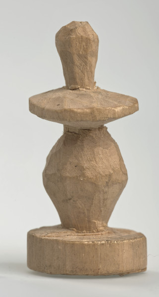 Ujęcie z boku [1]. Ze względu na swój okrągły kształt kolejne ujęcia z boku nie różnią się między sobą znacząco. Pionek został wykonany ręcznie z jednego kawałka jasnego drewna. Na całej powierzchni widoczne są ślady rzeźbienia przy pomocy noża. Obiekt w kolorze naturalnego drewna (nie został pomalowany, ani polakierowany). Od spodu widoczne oznaczenie składające się z dwóch przecinających się ze sobą linii („X”), które umożliwia podzielenie bierek pomiędzy dwóch graczy - wszystkie bierki są jednakowego koloru. Krawędź podstawy została dodatkowo obrysowana ołówkiem. Pionek składa się ze spłaszczonej podstawy w formie krążka, części środkowej w kształcie gruszki, spłaszczonego ronda, które zwęża się stopniowo ku górze oraz ku dołowi (przypominając dwa połączone ze sobą talerze, z których każdy skierowany jest dnem do zewnątrz) oraz zwieńczenia o prostej, rozszerzającej się ku górze formie (przypominającej pałkę).