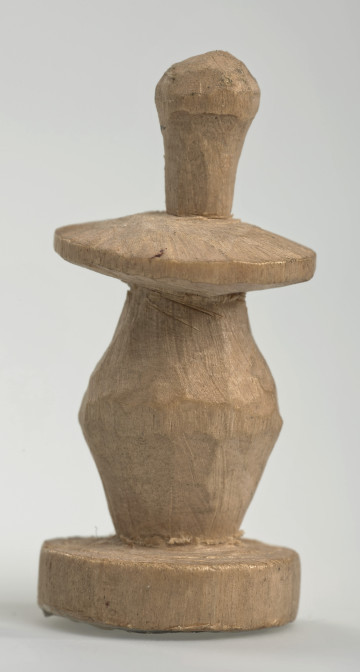 Ujęcie z boku [1]. Ze względu na swój okrągły kształt kolejne ujęcia z boku nie różnią się między sobą znacząco. Pionek został wykonany ręcznie z jednego kawałka jasnego drewna. Na całej powierzchni widoczne są ślady rzeźbienia przy pomocy noża. Obiekt w kolorze naturalnego drewna (nie został pomalowany, ani polakierowany). Krawędź podstawy została dodatkowo obrysowana ołówkiem. Pionek składa się ze spłaszczonej podstawy w formie krążka, części środkowej w kształcie gruszki, spłaszczonego ronda, które zwęża się stopniowo ku górze oraz ku dołowi (przypominając dwa połączone ze sobą talerze, z których każdy skierowany jest dnem do zewnątrz) oraz zwieńczenia o prostej, rozszerzającej się ku górze formie (przypominającej pałkę).