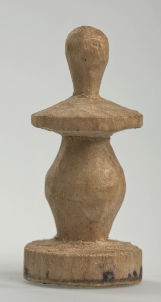 Ujęcie z boku [2]. Ze względu na swój okrągły kształt kolejne ujęcia z boku nie różnią się między sobą znacząco. Pionek został wykonany ręcznie z jednego kawałka jasnego drewna. Na całej powierzchni widoczne są ślady rzeźbienia przy pomocy noża. Obiekt w kolorze naturalnego drewna (nie został pomalowany, ani polakierowany). Krawędź podstawy została dodatkowo obrysowana ołówkiem. Pionek składa się ze spłaszczonej podstawy w formie krążka, części środkowej w kształcie gruszki, spłaszczonego ronda, które zwęża się stopniowo ku górze oraz ku dołowi (przypominając dwa połączone ze sobą talerze, z których każdy skierowany jest dnem do zewnątrz) oraz zwieńczenia o prostej, rozszerzającej się ku górze formie (przypominającej pałkę).