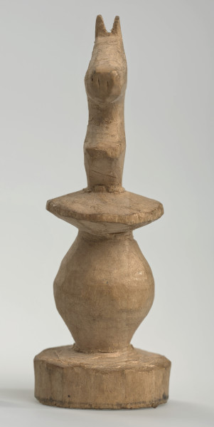 Ujęcie z przodu. 
Skoczek został wykonany ręcznie z jednego kawałka jasnego drewna. Na całej powierzchni widoczne są ślady rzeźbienia przy pomocy noża. Obiekt w kolorze naturalnego drewna (nie został pomalowany, ani polakierowany). Krawędź podstawy została dodatkowo obrysowana ołówkiem. Skoczek składa się ze spłaszczonej podstawy w formie krążka, części środkowej w formie walca rozszerzającego się po środku wysokości, spłaszczonego cylindrycznego elementu, który zwęża się stopniowo ku górze oraz ku dołowi (przypominając dwa połączone ze sobą talerze, z których każdy skierowany jest dnem do zewnątrz) oraz zwieńczenia w formie nawiązującej do głowy i szyi konia. Głowa oraz szyja są spłaszczone.