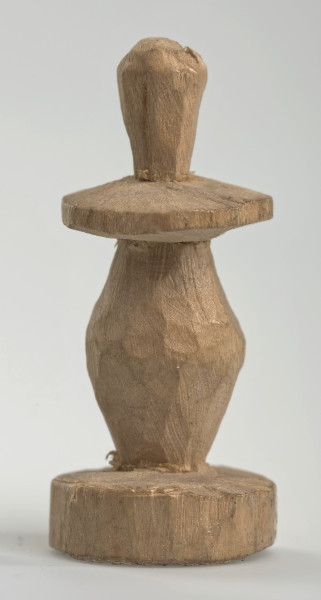 Ujęcie z boku [2]. Ze względu na swój okrągły kształt kolejne ujęcia z boku nie różnią się między sobą znacząco. Pionek został wykonany ręcznie z jednego kawałka jasnego drewna. Na całej powierzchni widoczne są ślady rzeźbienia przy pomocy noża. Obiekt w kolorze naturalnego drewna (nie został pomalowany, ani polakierowany). Od spodu widoczne oznaczenie składające się z dwóch przecinających się ze sobą linii („X”), które umożliwia podzielenie bierek pomiędzy dwóch graczy - wszystkie bierki są jednakowego koloru. Krawędź podstawy została dodatkowo obrysowana ołówkiem. Pionek składa się ze spłaszczonej podstawy w formie krążka, części środkowej w kształcie gruszki, spłaszczonego ronda, które zwęża się stopniowo ku górze oraz ku dołowi (przypominając dwa połączone ze sobą talerze, z których każdy skierowany jest dnem do zewnątrz) oraz zwieńczenia o prostej, rozszerzającej się ku górze formie (przypominającej pałkę).