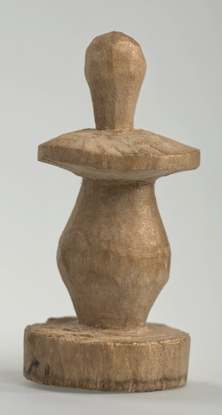 Ujęcie z boku [2]. Ze względu na swój okrągły kształt kolejne ujęcia z boku nie różnią się między sobą znacząco. Pionek został wykonany ręcznie z jednego kawałka jasnego drewna. Na całej powierzchni widoczne są ślady rzeźbienia przy pomocy noża. Obiekt w kolorze naturalnego drewna (nie został pomalowany, ani polakierowany). Krawędź podstawy została dodatkowo obrysowana ołówkiem. Pionek składa się ze spłaszczonej podstawy w formie krążka, części środkowej w kształcie gruszki, spłaszczonego ronda, które zwęża się stopniowo ku górze oraz ku dołowi (przypominając dwa połączone ze sobą talerze, z których każdy skierowany jest dnem do zewnątrz) oraz zwieńczenia o prostej, rozszerzającej się ku górze formie (przypominającej pałkę).