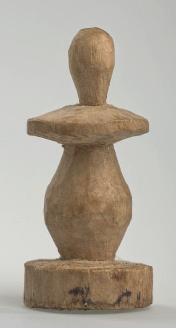 Ujęcie z boku [1]. Ze względu na swój okrągły kształt kolejne ujęcia z boku nie różnią się między sobą znacząco. Pionek został wykonany ręcznie z jednego kawałka jasnego drewna. Na całej powierzchni widoczne są ślady rzeźbienia przy pomocy noża. Obiekt w kolorze naturalnego drewna (nie został pomalowany, ani polakierowany). Krawędź podstawy została dodatkowo obrysowana ołówkiem. Pionek składa się ze spłaszczonej podstawy w formie krążka, części środkowej w kształcie gruszki, spłaszczonego ronda, które zwęża się stopniowo ku górze oraz ku dołowi (przypominając dwa połączone ze sobą talerze, z których każdy skierowany jest dnem do zewnątrz) oraz zwieńczenia o prostej, rozszerzającej się ku górze formie (przypominającej pałkę).