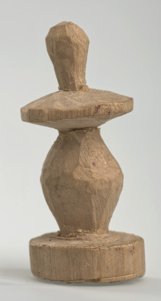 Ujęcie z boku [2]. Ze względu na swój okrągły kształt kolejne ujęcia z boku nie różnią się między sobą znacząco. Pionek został wykonany ręcznie z jednego kawałka jasnego drewna. Na całej powierzchni widoczne są ślady rzeźbienia przy pomocy noża. Obiekt w kolorze naturalnego drewna (nie został pomalowany, ani polakierowany). Od spodu widoczne oznaczenie składające się z dwóch przecinających się ze sobą linii („X”), które umożliwia podzielenie bierek pomiędzy dwóch graczy - wszystkie bierki są jednakowego koloru. Krawędź podstawy została dodatkowo obrysowana ołówkiem. Pionek składa się ze spłaszczonej podstawy w formie krążka, części środkowej w kształcie gruszki, spłaszczonego ronda, które zwęża się stopniowo ku górze oraz ku dołowi (przypominając dwa połączone ze sobą talerze, z których każdy skierowany jest dnem do zewnątrz) oraz zwieńczenia o prostej, rozszerzającej się ku górze formie (przypominającej pałkę).