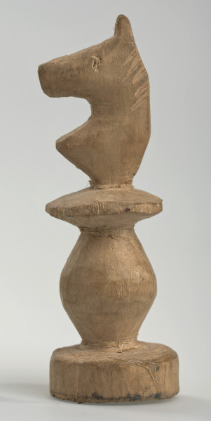 Ujęcie lewego boku. 
Skoczek został wykonany ręcznie z jednego kawałka jasnego drewna. Na całej powierzchni widoczne są ślady rzeźbienia przy pomocy noża. Obiekt w kolorze naturalnego drewna (nie został pomalowany, ani polakierowany). Krawędź podstawy została dodatkowo obrysowana ołówkiem. Skoczek składa się ze spłaszczonej podstawy w formie krążka, części środkowej w formie walca rozszerzającego się po środku wysokości, spłaszczonego cylindrycznego elementu, który zwęża się stopniowo ku górze oraz ku dołowi (przypominając dwa połączone ze sobą talerze, z których każdy skierowany jest dnem do zewnątrz) oraz zwieńczenia w formie nawiązującej do głowy i szyi konia. Głowa oraz szyja są spłaszczone. Widoczna wyżłobiona grzywa i oko.