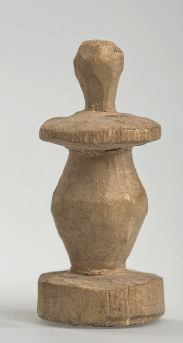 Ujęcie z boku [1]. Ze względu na swój okrągły kształt kolejne ujęcia z boku nie różnią się między sobą znacząco. Pionek został wykonany ręcznie z jednego kawałka jasnego drewna. Na całej powierzchni widoczne są ślady rzeźbienia przy pomocy noża. Obiekt w kolorze naturalnego drewna (nie został pomalowany, ani polakierowany). Od spodu widoczne oznaczenie składające się z dwóch przecinających się ze sobą linii („X”), które umożliwia podzielenie bierek pomiędzy dwóch graczy - wszystkie bierki są jednakowego koloru. Krawędź podstawy została dodatkowo obrysowana ołówkiem. Pionek składa się ze spłaszczonej podstawy w formie krążka, części środkowej w kształcie gruszki, spłaszczonego ronda, które zwęża się stopniowo ku górze oraz ku dołowi (przypominając dwa połączone ze sobą talerze, z których każdy skierowany jest dnem do zewnątrz) oraz zwieńczenia o prostej, rozszerzającej się ku górze formie (przypominającej pałkę).