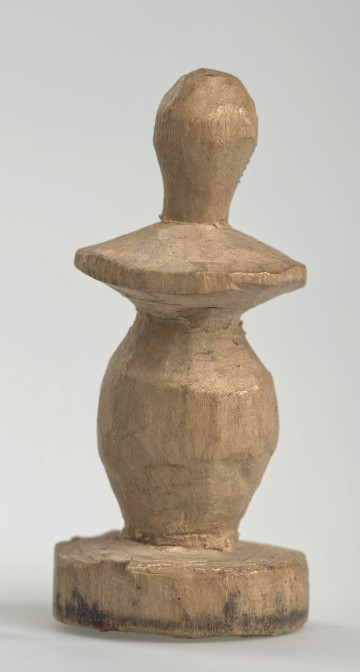 Ujęcie z boku [1]. Ze względu na swój okrągły kształt kolejne ujęcia z boku nie różnią się między sobą znacząco. Pionek został wykonany ręcznie z jednego kawałka jasnego drewna. Na całej powierzchni widoczne są ślady rzeźbienia przy pomocy noża. Obiekt w kolorze naturalnego drewna (nie został pomalowany, ani polakierowany). Od spodu widoczne oznaczenie składające się z dwóch przecinających się ze sobą linii („X”), które umożliwia podzielenie bierek pomiędzy dwóch graczy - wszystkie bierki są jednakowego koloru. Krawędź podstawy została dodatkowo obrysowana ołówkiem. Pionek składa się ze spłaszczonej podstawy w formie krążka, części środkowej w kształcie gruszki, spłaszczonego ronda, które zwęża się stopniowo ku górze oraz ku dołowi (przypominając dwa połączone ze sobą talerze, z których każdy skierowany jest dnem do zewnątrz) oraz zwieńczenia o prostej, rozszerzającej się ku górze formie (przypominającej pałkę).