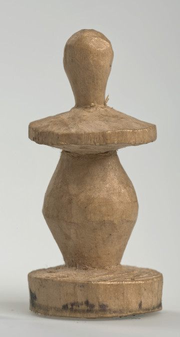 Ujęcie z boku [1]. Ze względu na swój okrągły kształt kolejne ujęcia z boku nie różnią się między sobą znacząco. Pionek został wykonany ręcznie z jednego kawałka jasnego drewna. Na całej powierzchni widoczne są ślady rzeźbienia przy pomocy noża. Obiekt w kolorze naturalnego drewna (nie został pomalowany, ani polakierowany). Krawędź podstawy została dodatkowo obrysowana ołówkiem. Pionek składa się ze spłaszczonej podstawy w formie krążka, części środkowej w kształcie gruszki, spłaszczonego ronda, które zwęża się stopniowo ku górze oraz ku dołowi (przypominając dwa połączone ze sobą talerze, z których każdy skierowany jest dnem do zewnątrz) oraz zwieńczenia o prostej, rozszerzającej się ku górze formie (przypominającej pałkę).