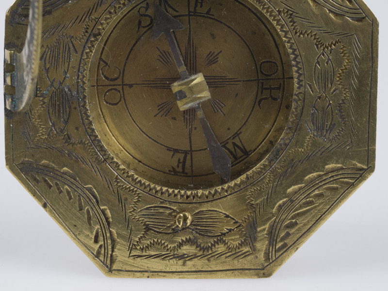 zbliżenie na ośmiokątną, grawerowaną w motywy geometryczno-floralne tarczę zegara z przeszkloną mosiężną puszką z kompasem w części centralnej (szkiełko otwarte)