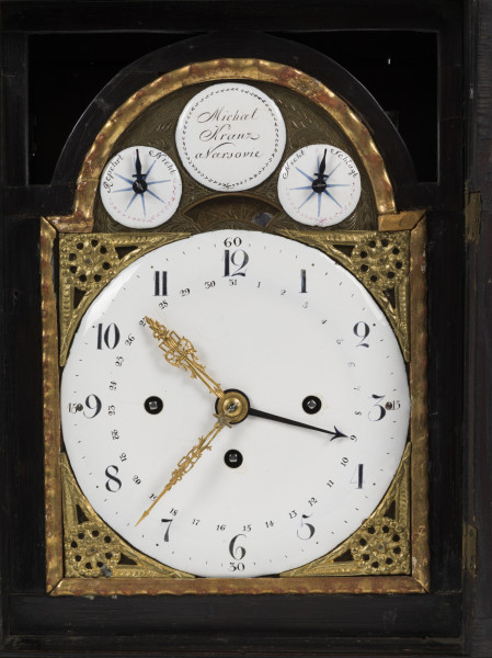 zbliżenie na tarczę zegara ze wskazaniami kalendarza, dni miesiąca, godzinami, kwadransami, minutami i sekundami, z repetierem oraz drewnianą, przeszkloną obudowę z dekoracyjnymi ażurami akustycznymi z mosiądzu grawerowanego