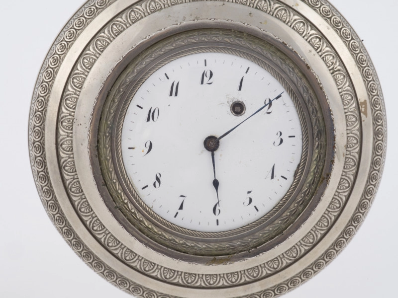 zbliżenie na porcelanową okrągłą tarczę w oprawie z białego metalu, dekorowaną obiegającymi w koło pasami na przemian gładkimi i pokrytymi plastyczną ornamentacją roślinną