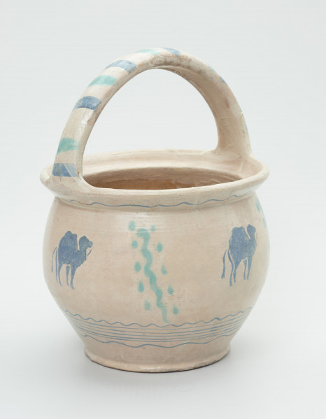 garnek z kabłąkowym uchem - Ujęcie z boku. Garnek uformowany na kole garncarskim z gliny o jasnej barwie (z nieznaczną ilością tlenków żelaza). Dno lekko zaznaczone przechodzi w pękaty brzusiec zakończony wlewem z kołnierzem wygiętym mocno na zewnątrz. Do krawędzi wlewu przymocowane taśmowe ucho. Szkliwo w tonacji szarej na powierzchni licowej i wewnątrz naczynia. Malatura wykonana naszkliwnie - odręcznie oraz stemplem przedstawiającym wielbłąda. Brzusiec w dolnej partii i pod kołnierzem zdobiony liniami falistymi. Jego środkowa część zdobiona pionową linią falistą z kropkami po obu jej stronach naprzemiennie ze stemplem z wielbłądem. Każdy z motywów powtórzony po cztery razy. Ucho zdobione grubymi kreskami. Całość malatury w kolorach niebieskim i zielonym .
