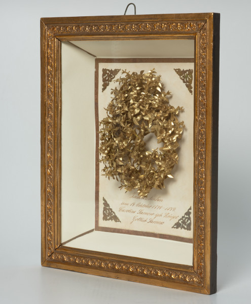 naczynie zasobowe - Ujęcie z przodu skosem w lewo. Wieniec w ramie za szkłem. Rama  głęboka, wewnątrz wyłożona białym papierem. Listwy ozdobne, złocone. Wewnątrz centralnie umieszczony, wykonany z pozłacanego papieru modelowany na koronę wieniec składający się z drobnych listków i pięciopłatkowych kwiatków stylizowanych na mirt. Poniżej niego odręczny napis w języku niemieckim. W rogach przyklejone trójkątne ozdoby – stylizowane motywy roślinne. Całość ujęta w obwódkę wykonaną z papierowych pasków z wytłaczanym, delikatnym wzorkiem. Cieńsze paski, bez zdobień, przyklejone są w rogach ramy.