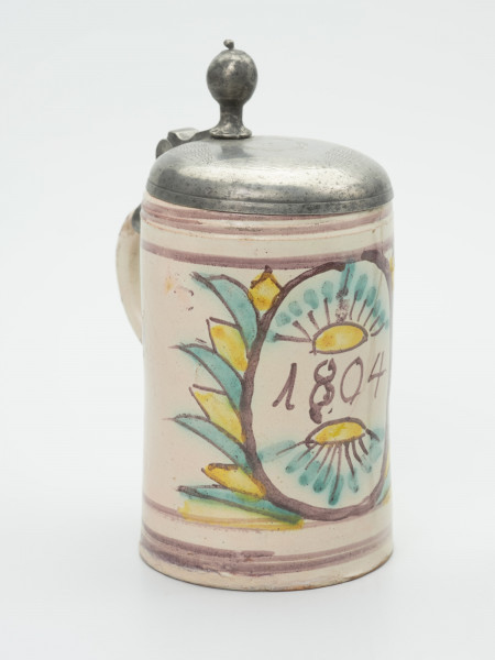 kufel - Ujęcie z przodu skosem w prawą stronę. Naczynie gliniane uformowane na kole garncarskim. Kształt cylindryczny. Pod krawędzią szerokiego wlewu dwa dookolne ryte paski. Poniżej zamocowane taśmowe ucho sięgające poniżej połowy wysokości kufla. Na uchu mocowana cynowa pokrywka ozdobnie grawerowana, z paluchem w kształcie kuli. Szkliwiony obustronnie, dekorowany ręcznie. Dominantą kompozycji jest owalny kartusz - kwiat zdobiony na zewnątrz i w środku stylizowanymi motywem kwiatowymi oraz datą (1804) w centrum. Bliżej ucha naczynia także kwiaty. Całość w kolorach: zielonym, żółtym i fioletowym .