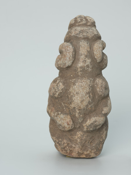 rzeźba; Figura kultu zmarłych - Ujęcie z tyłu. Rzeźbiona w szarobeżowym steatycie siedząca postać ludzka z dużą głową o masywnych, okragłych okularach i przykurczonymi nogami, których kolana sięgają brody.