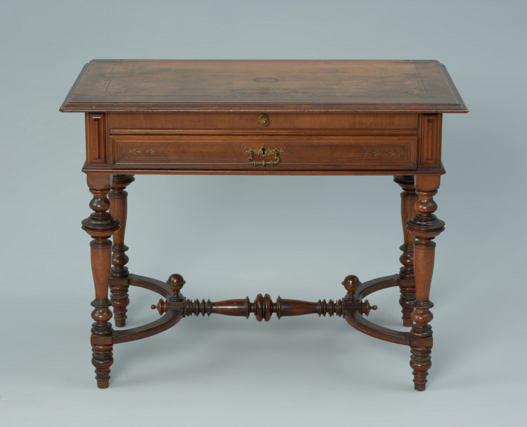 Drewniany stolik z profilowanymi nożkami - Ujęcie z przodu. Stolik z drewna fornirowanego na czterech toczonych, profilowanych nogach. Blat o profilowanych krawędziach, pokryty rytym ornamentem; w tylnej części blatu dwa otwory, świadczące o pierwotnym wyposażeniu biurka w nadstawkę o charakterze sekretarzyka. Pod blatem zakończona poziomym profilem skrzynia mieszcząca wysuwany blat i szufladę. Nogi tralkowe, połączone w dolnej części poziomą konstrukcją przypominającą kształtem podwójne Y, utworzoną przez dwie łukowo wygięte listwy łączące parami boczne nogi oraz przecinającą półkola ozdobną tralkę. W miejscach skrzyżowania poprzeczki z łukami okazałe toczone gałki skierowane ku górze oraz mniejsze poziome w przedłużeniach poprzecznej tralki. Po obu stronach szuflady i wysuwanego blatu pionowe, prostokątne kaboszony. Czoło szuflady płycinowe, z profilowanym obramieniem, ozdobione poziomym pasem rytego ornamentu geometryczno-roślinnego. W centrum mosiężny uchwyt na zawiasie oraz rozetkowa oprawa dziurki od klucza zamka (ukrytego wewnątrz szuflady); uchwyt ozdobiony ornamentem zwijanym i roślinnym. Wysuwany blat o profilowanej krawędzi zewnętrznej, wyłożony tkaniną; po wysunięciu jego czoło na zawiasach odchyla się w dół. Do wyciągania blatu służy umieszczona centralnie mosiężna gałka. Głównym motywem rytej dekoracji blatu biurka jest płomienista waza wśród wici roślinnej. W zagłębieniach rytów oraz na niektórych profilach ślady złoceń.