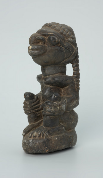 rzeźba; przedmiot obrzędowy; Figura kultu sił wegetacji - Ujęcie z przodu z lewej strony. Rzeźbiony w grafitowym steatycie, na owalnej podstawie, mężczyzna w pozycji siedzącej z opaską przedstawioną za pomocą wypukłego ornamentu kostkowego, której końce opadają na ramiona i plecy postaci.
