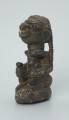 rzeźba; przedmiot obrzędowy; Figura kultu sił wegetacji - Ujęcie z przodu z lewej strony. Rzeźbiony w grafitowym steatycie, na owalnej podstawie, mężczyzna w pozycji siedzącej z opaską przedstawioną za pomocą wypukłego ornamentu kostkowego, której końce opadają na ramiona i plecy postaci.
