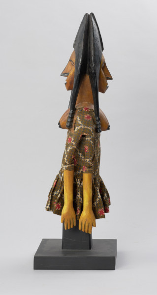 lalka teatralna: Djinè Faro (bóstwo Faro) - Ujęcie z boku z lewej strony. Drewniana lalka przedstawiająca postać kobiecą o jasnej cerze. Twarz pociągła, nos długi, duże usta, ozdobne nakrycie głowy w kolorze czarnym. Ubrana we wzorzystą sukienkę odsłaniającą nagie piersi.